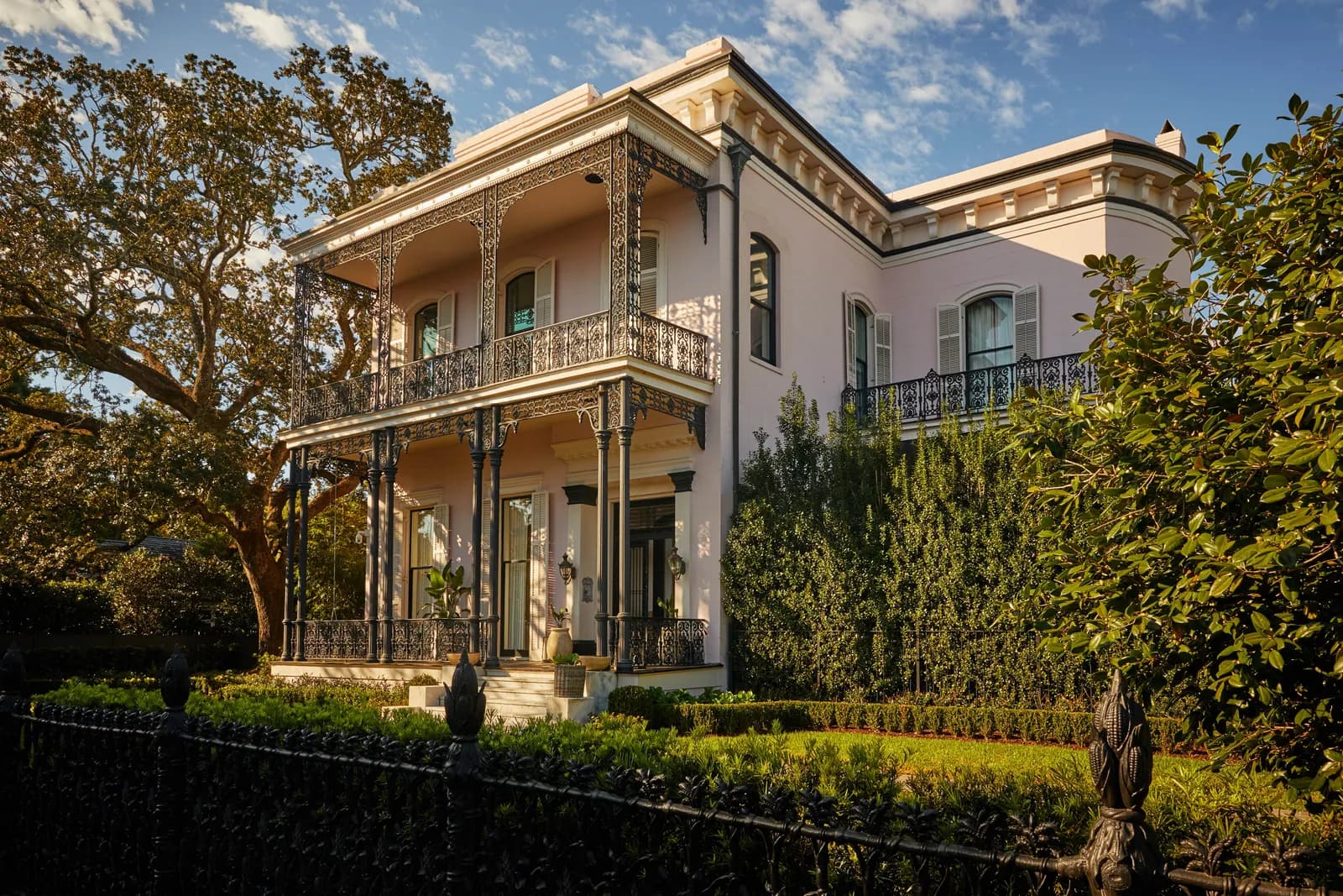 A pink Garden District mansion at golden hour, its double galleries wrapped in ornate cast iron, behind a cornstalk fence and live oaks